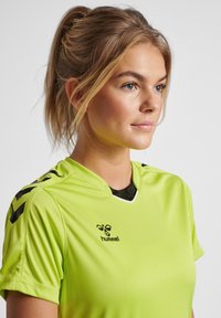 Grøn atletisk trøje lavet af glat stof med v-hals, sorte accenter på skuldrene og et logo på brystet. Let tekstur.