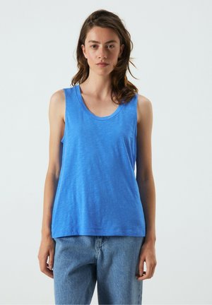 Giovane donna che indossa una canotta blu senza maniche e jeans azzurri, in piedi e rivolta in avanti su uno sfondo chiaro.