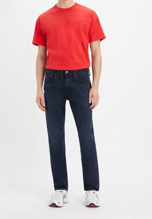 Mand iført rød kortærmet T-shirt, mørkeblå jeans og hvide sneakers, stående med arme afslappet langs siden på ensfarvet hvid baggrund.