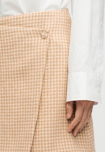 Μπεζ και λευκή φούστα με σχέδιο houndstooth και μεγάλο κουμπί, φορεμένη με λευκό πουκάμισο μακρυμάνικο και χέρι που ακουμπά στην πλευρά.