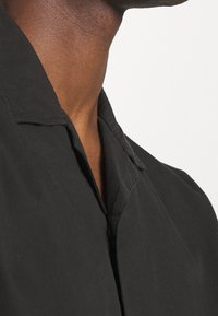 Černá košile s krátkým límečkem a měkkou texturou, s knoflíkovým zapínáním. Viditelný steh dodává designu jemný detail.