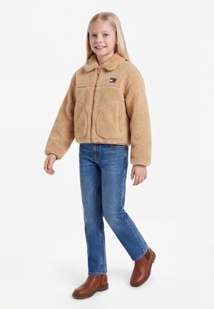 Lyshåret pige iført en beige fleecejakke, blå jeans og brune ankelstøvler, står med det ene ben let løftet og smiler.