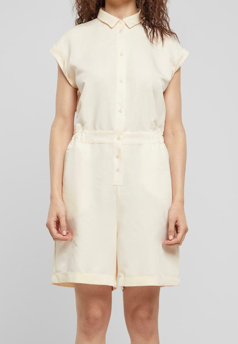 Femme portant une combinaison courte crème à manches courtes, boutonnée avec col et taille élastique, debout devant un fond gris uni.