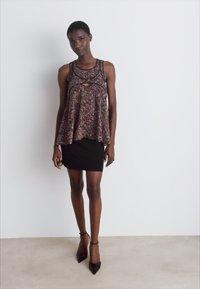 Haut sans manches à motifs dans des teintes sombres avec des accents en dentelle, associé à une jupe noire ajustée. Le modèle se tient sur un fond clair.
