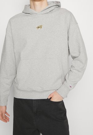 Person trägt hellgrauen Kapuzenpullover mit kleinem besticktem Logo auf Brust und Ärmel, kombiniert mit schwarzen Hosen.