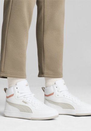 Personne portant des baskets montantes blanches Puma avec des accents beiges, des chaussettes blanches et un pantalon beige sur un fond neutre.