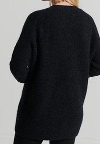 Svart tröja gjord av mjukt, texturerat tyg med en avslappnad passform, med rundad halsringning och långa ärmar som sträcker sig förbi höfter.