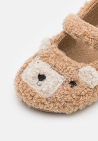 Miękka, beżowa skórzana bucik dla malucha z kręconą fakturą i wzorem twarzy misia na palcu, z białymi uszami oraz czarnymi oczami i nosem.