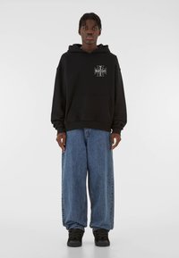 Black hooded sweatshirt with front pocket and graphic design on chest, paired with loose-fitting blue denim jeans and black sneakers.