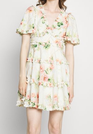 Woman wearing short white floral dress with ruffled tiers and puffed sleeves, standing against plain light background.