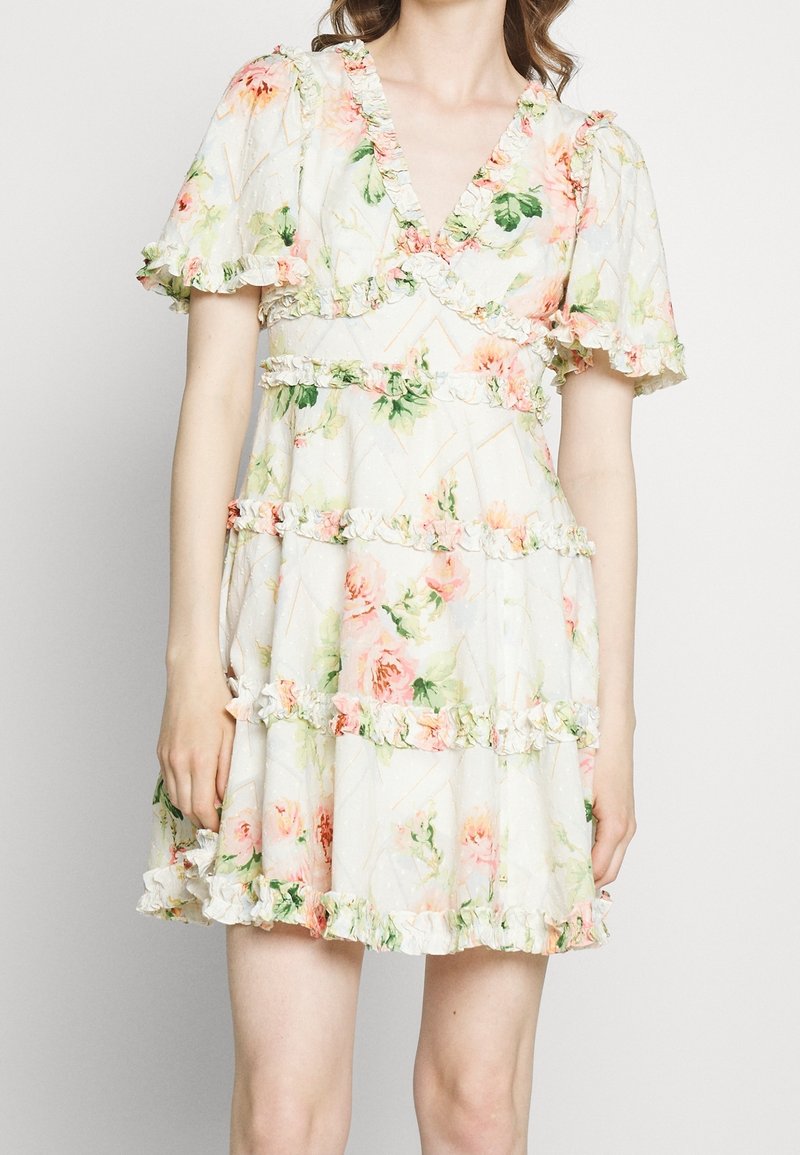 Femme portant une courte robe blanche à fleurs avec des volants superposés et des manches bouffantes, debout devant un fond clair uni.