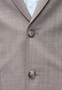 Γκρι υφασμάτινο σακάκι με δύο μεγάλα σκούρα κουμπιά πάνω από ένα ανοιχτό γαλάζιο πουκάμισο με γιακά που φαίνεται από κάτω.