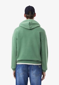 Grüne Kapuzen sweatshirt mit entspannter Passform, weichem Material und einer Kängurutasche vorne; kombiniert mit blauen Jeans.