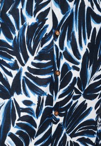 Πουκάμισο με κουμπιά, με μεγάλα τροπικά σχέδια φύλλων σε σκούρο και ανοιχτό μπλε χρώμα πάνω σε λευκό φόντο.