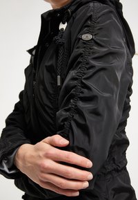 Veste imperméable noire avec capuche, détails de tissu froncé sur les manches et cordon texturé. Accents en métal visibles.