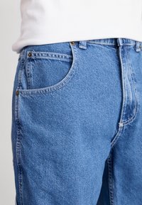 Vaqueros de mezclilla azul claro con un corte clásico, que cuentan con herrajes metálicos y costuras visibles. Bolsillos visibles en la parte delantera y trasera.