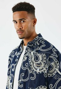 Giacca di jeans di colore blu scuro, con ricami bianchi a paisley, chiusura con bottoni e taschino sul petto con etichetta.