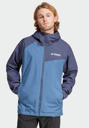 Veste imperméable bleue et marine foncé avec une fermeture éclair, capuche et logo TERREX. Elle présente une texture lisse et légère avec des blocs de couleur contrastants.