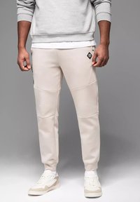 Světlé béžové tepláky s hladkou texturou, úzkým střihem a elastickými lemy. Obsahují boční kapsu s detailem černé nášivky.