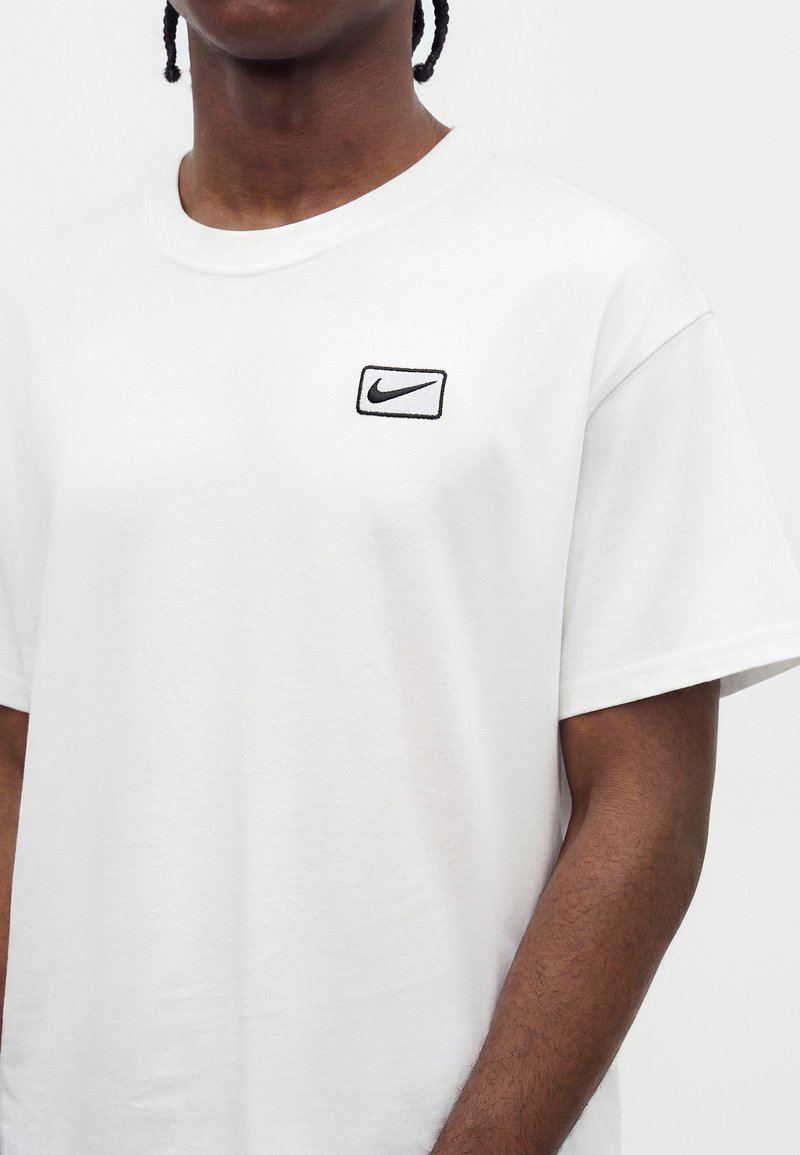 Mann trägt ein schlichtes weißes Nike-T-Shirt mit einem kleinen schwarzen Nike-Logo auf der linken Brust, vor weißem Hintergrund.