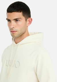 Lys beige hoodie med snor, blød tekstur, med et diskret prægget logo. Enkel design uden mønstre eller yderligere accenter.