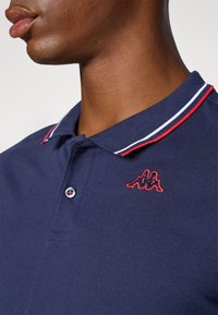 Marine polo skjorte med kontrastfarget rød og hvit stripet krage, to knapper, og en rød brodert logo på brystet. Glatt bomullstekstur.