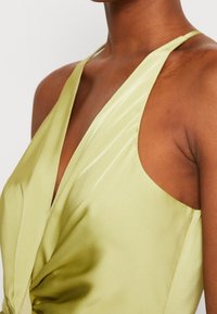 Zelené saténové šaty s hlubokým výstřihem, halterovým zapínáním a zkroucenými detaily v pase. Hladká textura s lesklým povrchem.