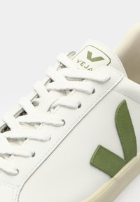 Hvite Veja-sneakers med grønn logo, med teksturerte lisser og en glatt skinnoverdel. Myk beige såle kontrasterer med overdelens design.