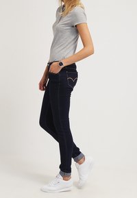 Mörkblå slim-fit jeans med ett hjärtformat sömnadsdetalj på bakfickorna, kombinerat med en grå pikétröja och vita sneakers.