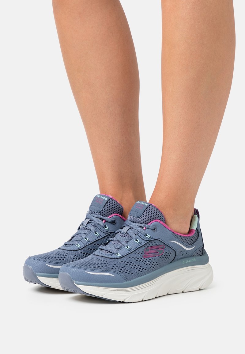 Jambes portant des baskets Skechers en mesh gris et rose avec des semelles blanches, debout sur un fond clair uni.