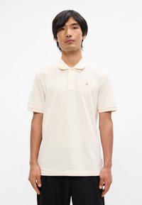 Κ ρέμα πόλο μπλούζα με κλασικό γιακά, κοντά μανίκια και τρία κουμπιά, με μικρό χρυσό λογότυπο στην αριστερή πλευρά του στήθους.