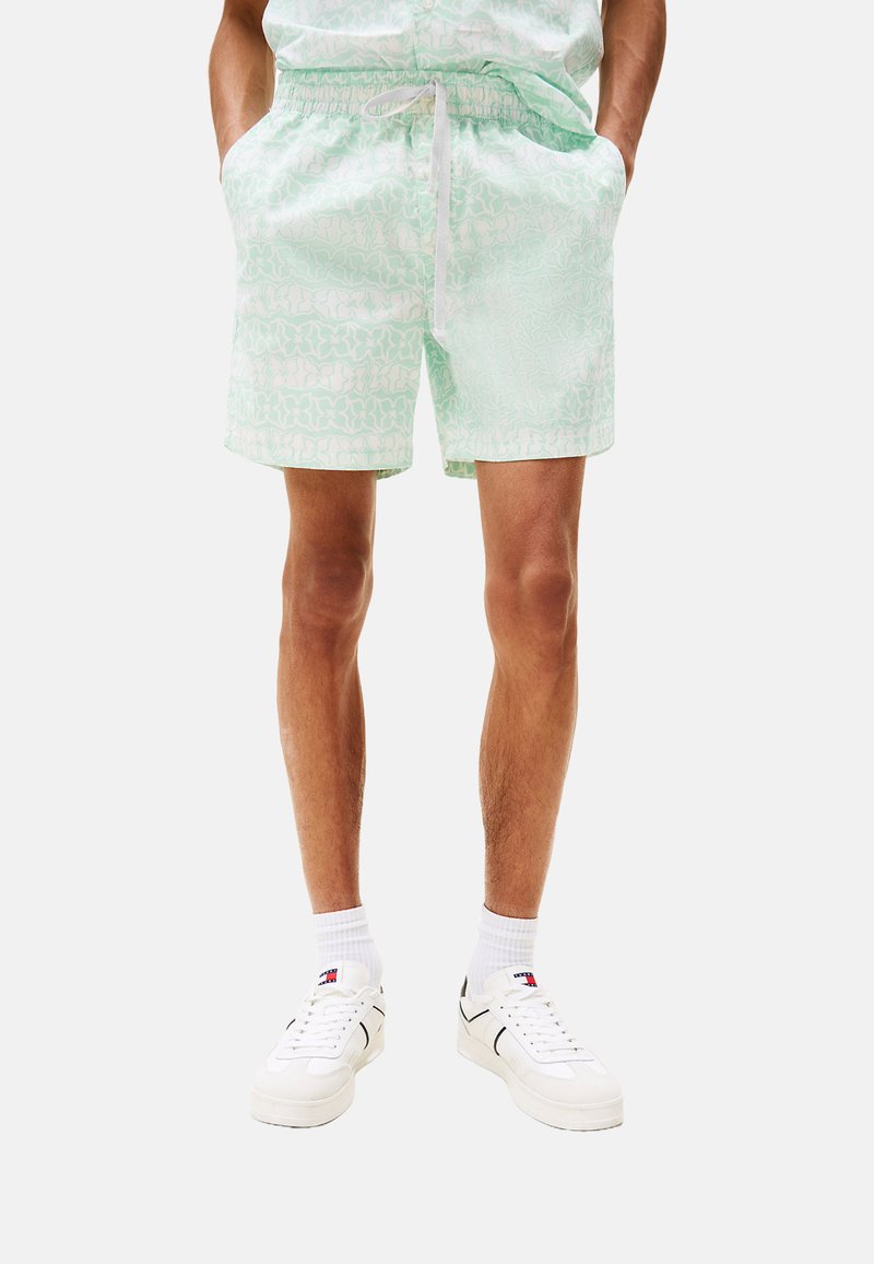 Pantaloni da bagno verde chiaro con motivo, dotati di vita elasticizzata e coulisse. Abbinati a scarpe da ginnastica bianche e calze bianche.