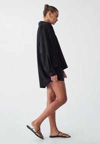 Černá oversized košile s límečkem, knoflíkovými manžetami a bočními rozparky. Nosit s černými sandály na popruhy. Hladká textura.