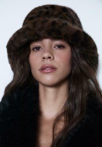 Cappello a secchiello a stampa leopardo marrone realizzato in morbido pelliccia, con una larga visiera soffice. Indossato con un cappotto di pelliccia nero.