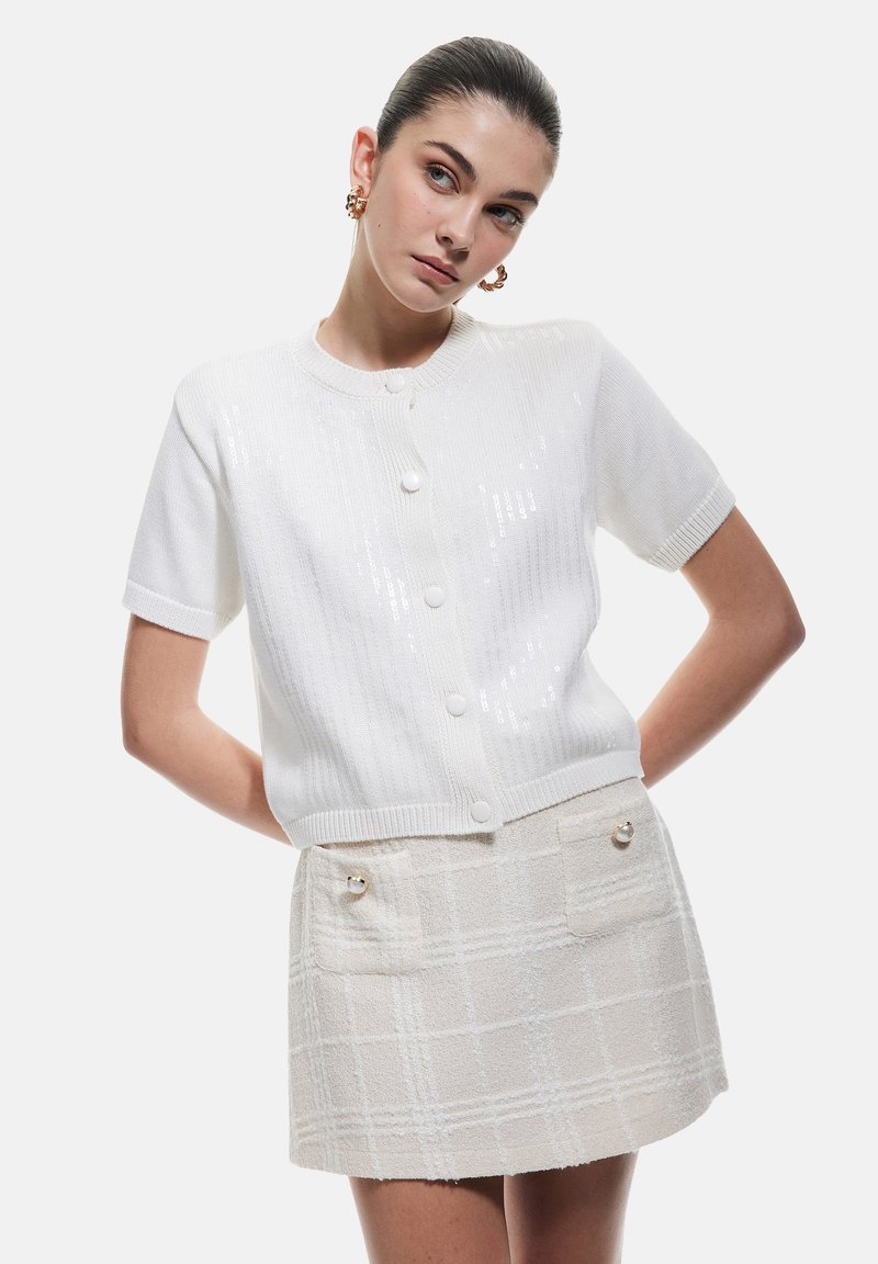 Cardigan bianco lavorato a maglia con maniche corte, caratterizzato da una chiusura con bottoni e una leggera lucentezza. Abbinato a una minigonna testurizzata beige chiaro.
