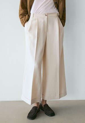 Pantalon beige large raccourci avec plis avant, porté avec des chaussures noires à enfiler et une veste marron sur un haut blanc. Mains dans les poches.