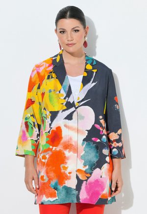 Vrouw met donker haar die grote rode en gouden oorbellen draagt en een kleurrijke blazer met bloemenpatroon en felrode broek, staand tegen een witte achtergrond.