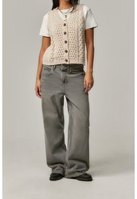 Béžová pletená vesta s texturovaným vzorem, zapínání na knoflíky, nošena přes bílý tričko. Doplněno volnými šedými džíny a černými botami.