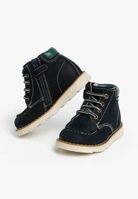 Bottines en cuir bleu marine avec des coutures blanches, semelles en caoutchouc et accents verts. Elles possèdent des lacets et une fermeture éclair sur le côté pour un enfilage facile.