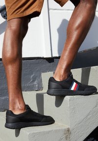 Personne portant des baskets noires avec des bandes rouges, blanches et bleues montant des escaliers en béton gris à l'extérieur.
