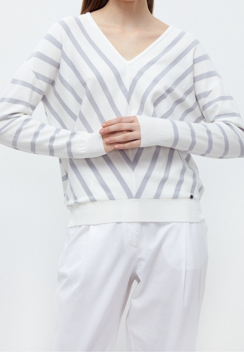 Femme portant un pull blanc à col en V avec des rayures diagonales grises et un pantalon blanc, mains croisées au niveau du ventre sur un fond uni.
