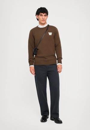 Bruine gebreide trui met een wit letter "W" logo, gedragen over een witte blouse met kraag, gecombineerd met donkere marineblauwe broek en zwarte schoenen.