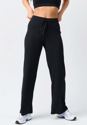 Frau trägt schwarze, breitbeinige, gerippte Hose und weiße Sportschuhe, steht mit Händen an der Taille und passt diese an.