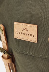Olivengrønn lerretsveske med et brunt lærfang som har påskriften "DOUGHNUT" i relieff, sikret med en lærstropp og metalldetaljer.
