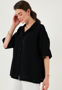 Black, collared shirt with short sleeves, featuring a textured fabric, button front, and a relaxed fit. Worn with gold-tone jewelry.