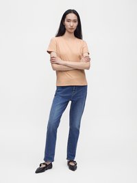 Femme aux longs cheveux noirs debout, bras croisés, portant un t-shirt beige, un jean bleu et des chaussures plates pointues noires sur fond blanc.