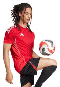 Maillot de sport rouge avec des accents blancs, associé à un short noir et des chaussettes noires à mi-mollet, mettant en scène un ballon de football en mouvement.