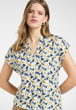 Kvinde iført kortærmet bluse med blå, sort og brun abstrakt fjerlignende mønster, hænder på hofterne, neutral baggrund.