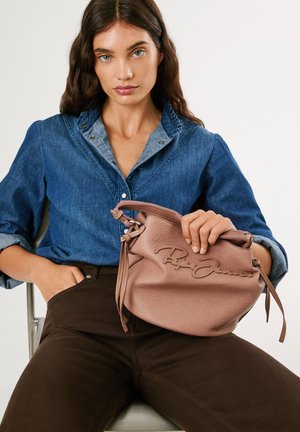 Ung kvinna med långt mörkt hår i blå jeansskjorta och mörka byxor sitter och håller en strukturad mauve läderhandväska med en broderad logotyp.