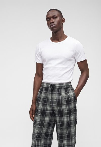 Jeune homme portant une chemise blanche unie à manches courtes et un pantalon à carreaux noir et blanc, debout avec une main dans la poche.